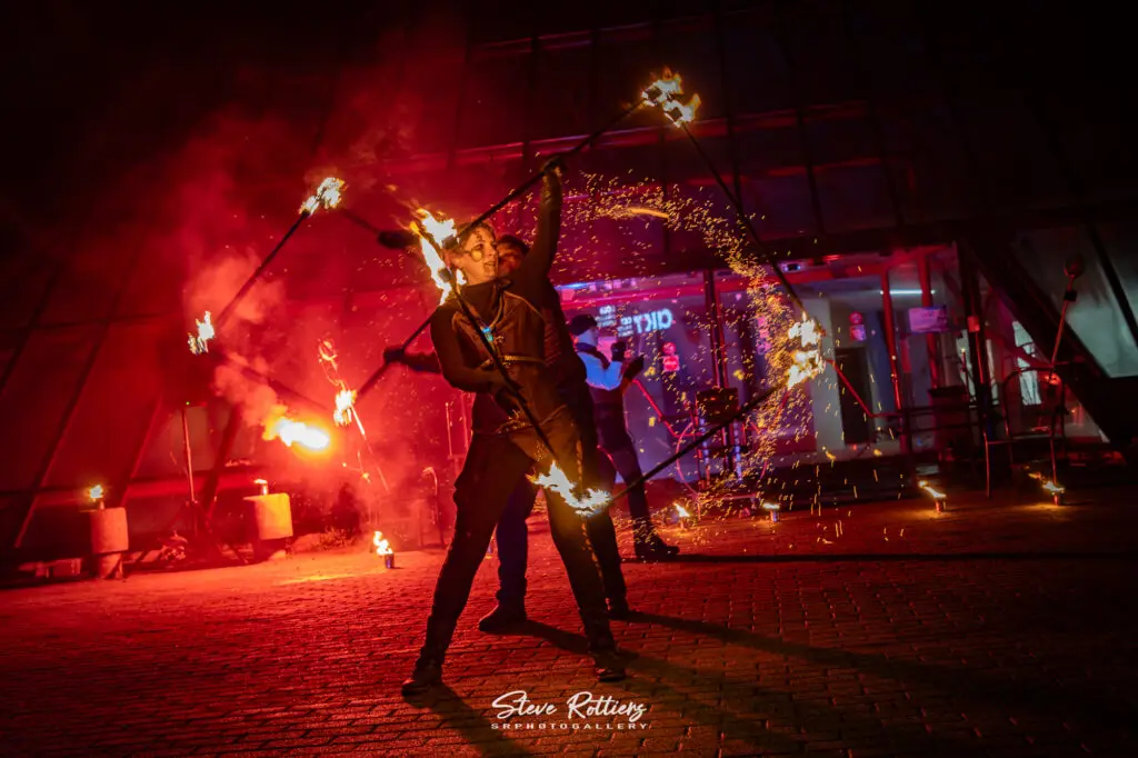 Deux jongleurs de feu exécutant une chorégraphie millimétrée avec un instrument unique dans le spectacle Étincelle Mécanique, lors d’un événement en province du Hainaut.
